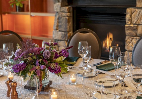 A decorated dining table with a floral centerpiece, candles, wine glasses, and silverware in front of a fireplace.