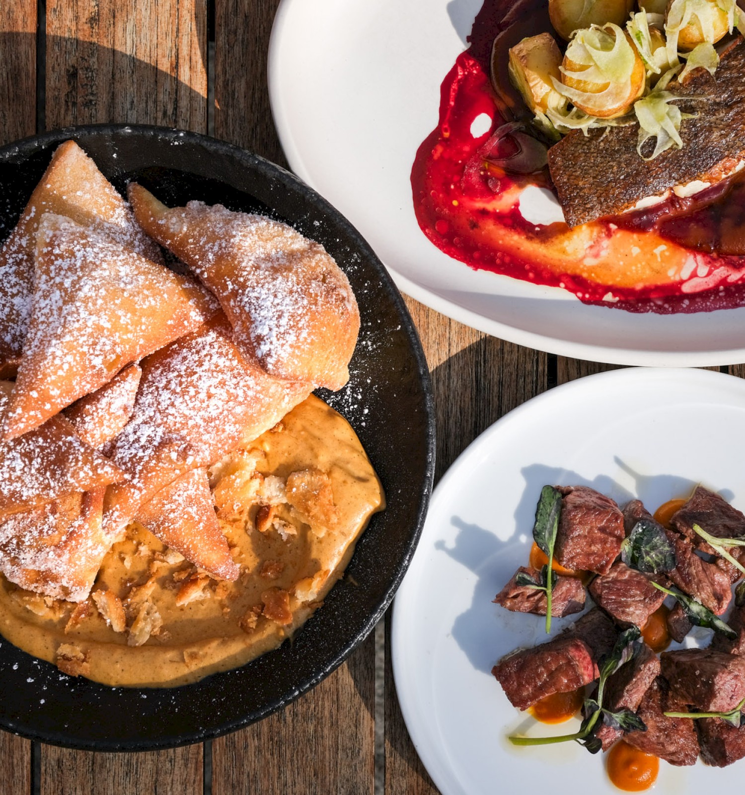 Three dishes of gourmet food are arranged on wooden surface: fried pastries with powdered sugar, fish with sauce, and beef with vegetables.
