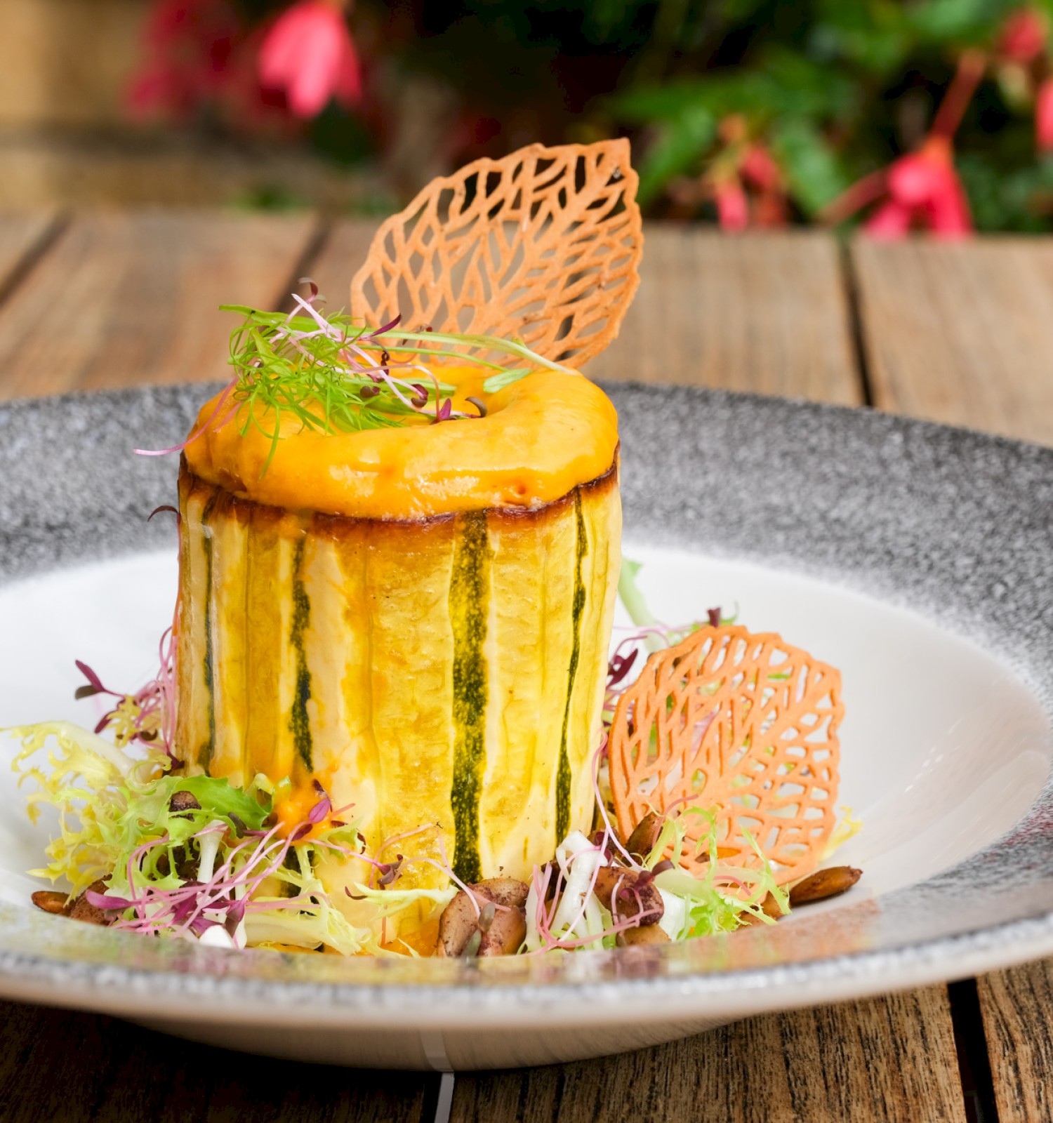 A gourmet dish featuring a cylindrical container topped with a yellow sauce, garnished with microgreens and orange lacy decorations.