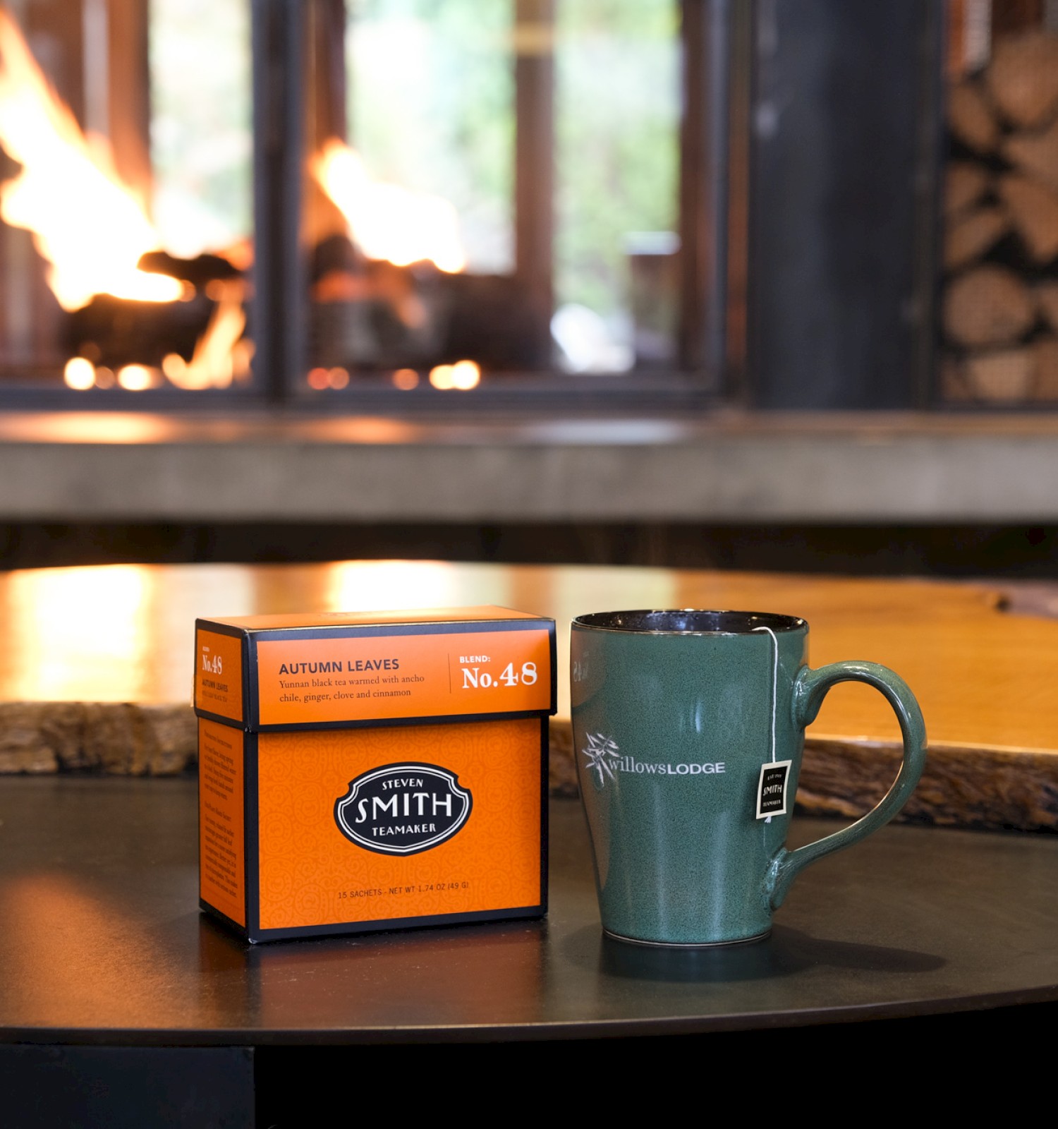 A cozy scene with a box of autumn tea and a green mug on a table in front of a fireplace with logs and flames.