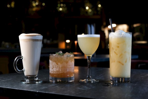 Four different cocktails are shown on a dark bar counter, each with unique glassware and garnishes, in a dimly lit bar setting.