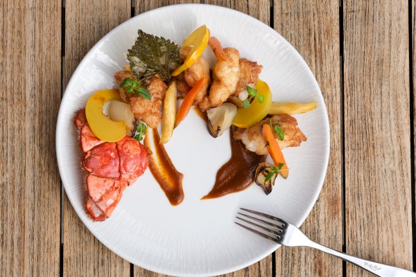 A seafood dish served with vegetables and sauces on a white plate placed on a wooden table.