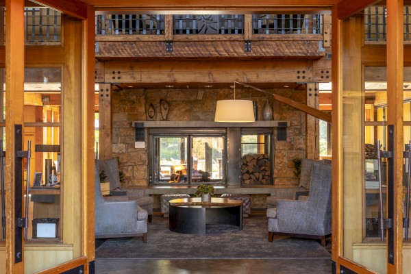 A cozy living room with gray armchairs, a wooden coffee table, a stone fireplace, and a wood-paneled ceiling in a rustic lodge-style setting.