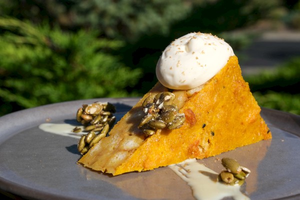 A slice of pumpkin pie topped with whipped cream, pumpkin seeds, and drizzled with cream on a gray plate outdoors.