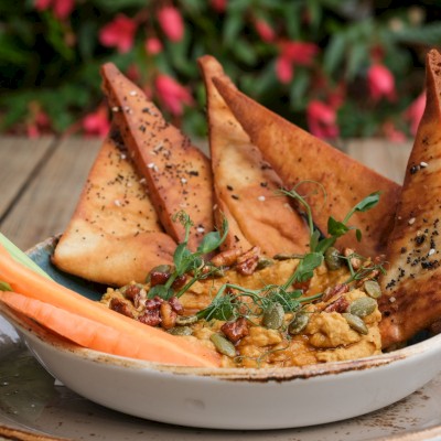 A bowl of Indian-style hummus topped with crispy papadum, garnished with greens, seeds, and vegetable sticks on a wooden table.