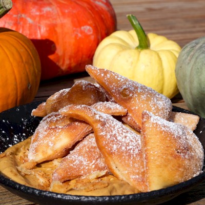 Fried pumpkin slices dusted with powdered sugar are served in a black dish, surrounded by colorful pumpkins on a wooden surface.