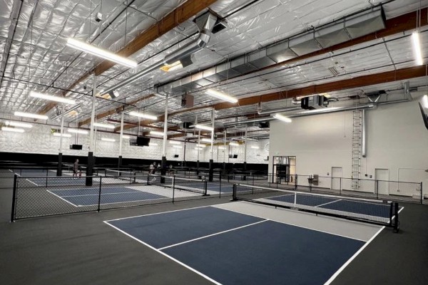 An indoor tennis facility with multiple courts, high ceiling with exposed pipes, and several people playing.