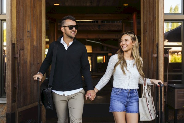 A couple is walking hand in hand, smiling, with luggage, exiting a building, enjoying a sunny day.