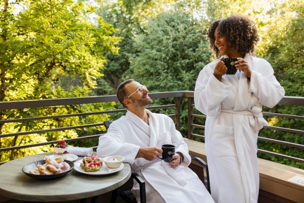 Two people in robes enjoying coffee on a balcony with fruit and pastries, surrounded by lush greenery.