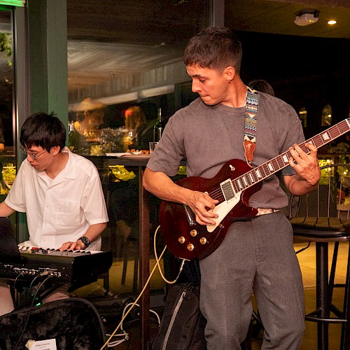 Musicians perform live jazz music at an indoor event, with one playing the keyboard and the other on an electric guitar.