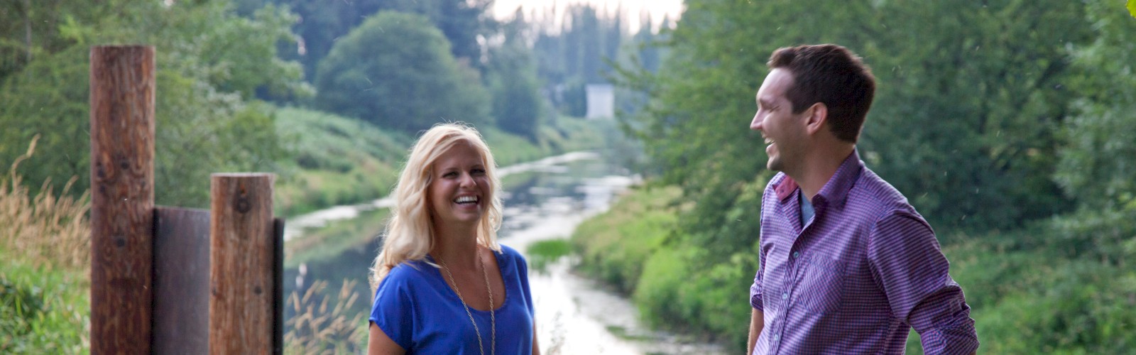 Two people stand with bicycles by a river in a lush, green setting, smiling and engaged in conversation.