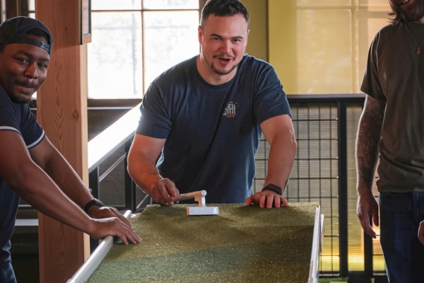 Three men are enjoying a game of shuffleboard indoors, smiling and having a good time together.