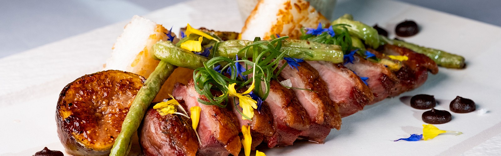 A beautifully plated gourmet steak dish with vegetables, edible flowers, sauce, and a ceramic jug on a white plate.