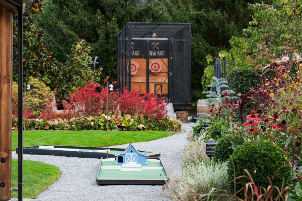 A backyard with a mini golf course, a wooden shed, athletic targets, and lush colorful plants and trees.