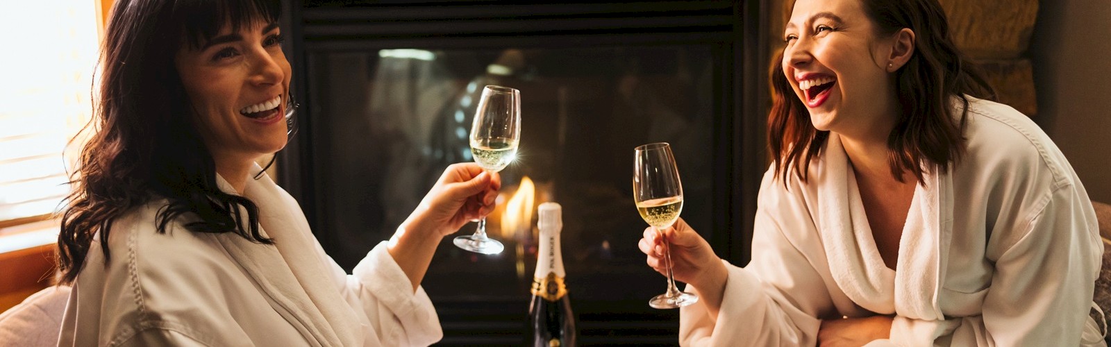 Two people in robes are sitting by a fireplace, smiling and clinking wine glasses, with a champagne bottle on a small wooden table.