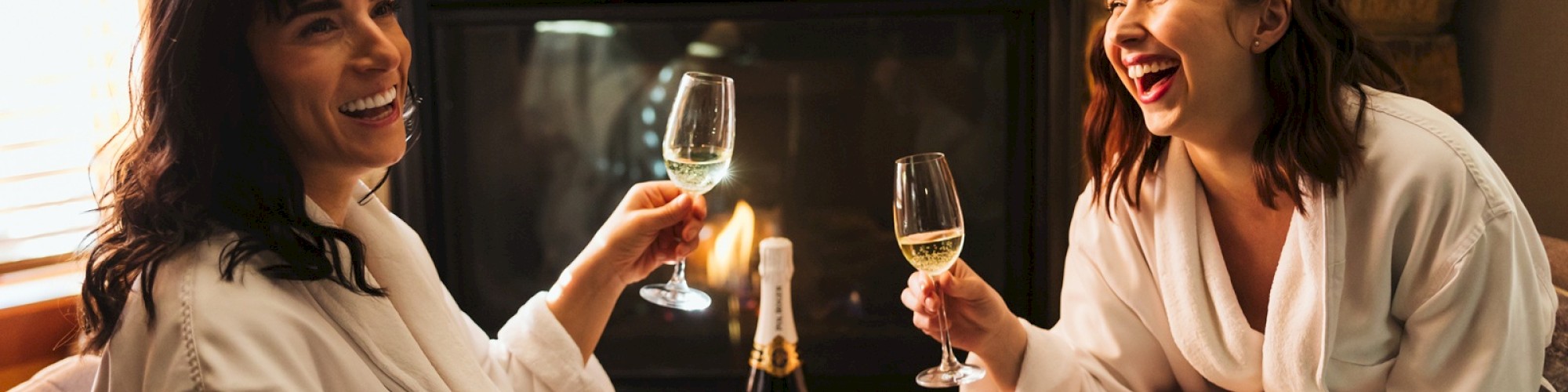 Two people in robes are sitting by a fireplace, smiling and clinking wine glasses, with a champagne bottle on a small wooden table.