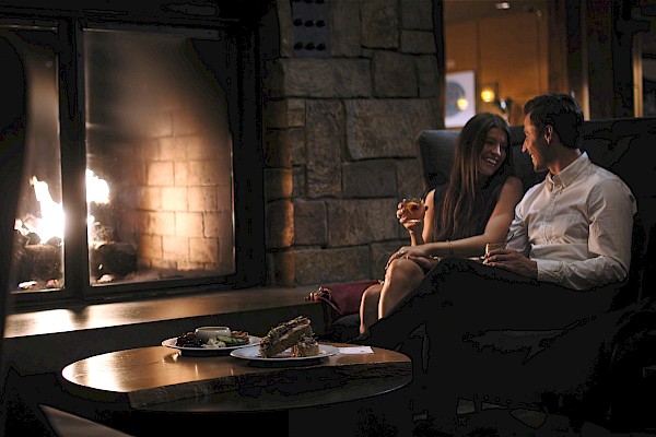 A couple enjoys drinks by a fireplace in a cozy, dimly lit restaurant or lounge setting.
