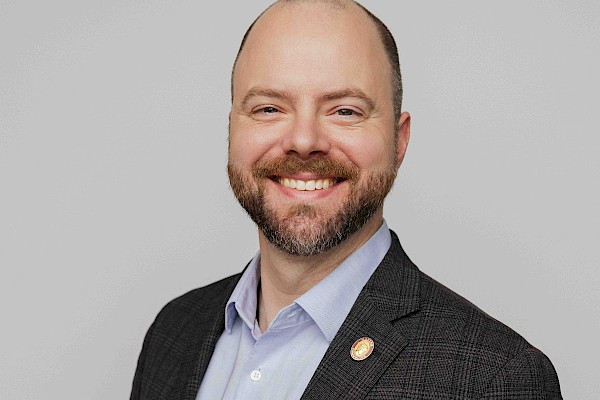 A smiling man in a dark blazer and light blue shirt, posing for a professional portrait with a neutral gray background.