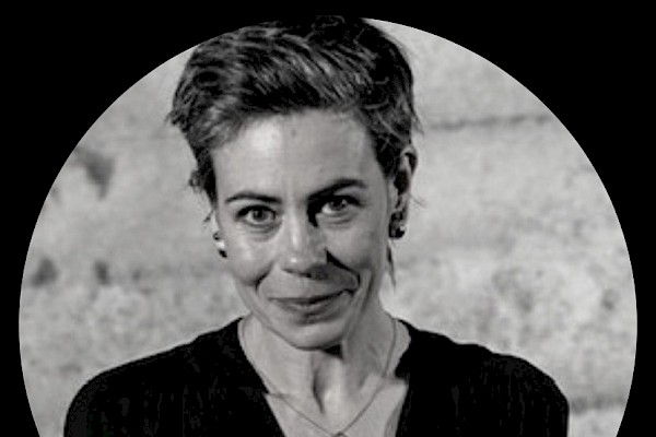 A grayscale portrait of a woman with short hair, blazer, and necklace, centered within a circular frame against a dark background.