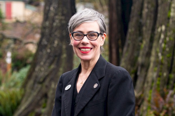 A smiling woman with short gray hair and glasses, wearing a black blazer, standing outdoors among trees.