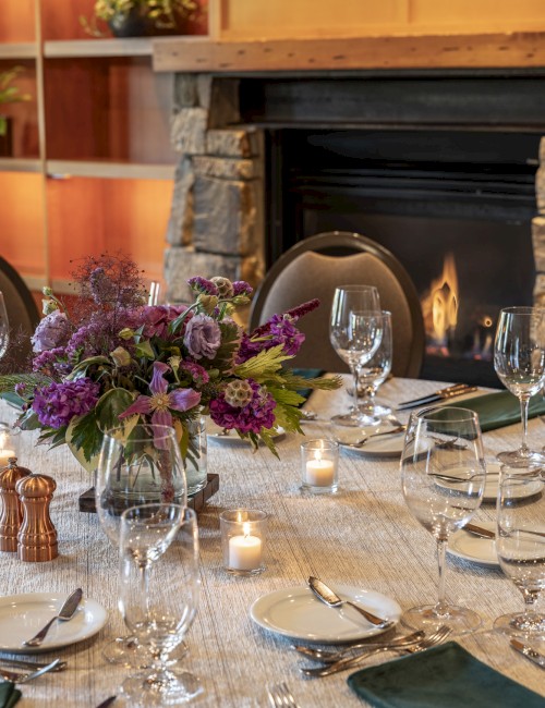 A beautifully set dining table with flowers, candles, glasses, and cutlery, near a cozy fireplace in a warmly decorated room.