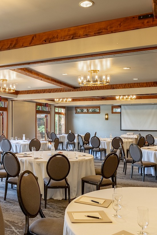 A large, well-lit banquet room set up with round tables, glasses, notepads, and a projector screen, ready for a conference or event.