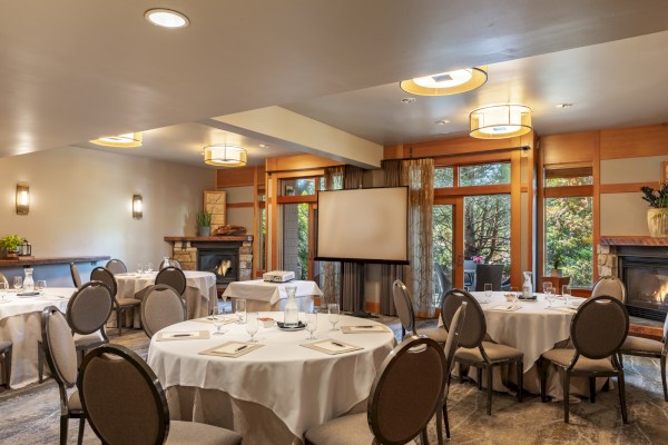 A conference room with round tables set for a meeting, featuring a projector screen, fireplace, and large windows showing trees.