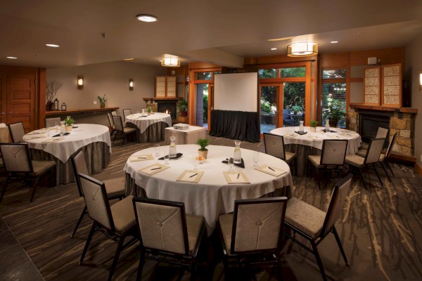 A conference room is set up with round tables, chairs, a projector screen, and notepads, ready for a meeting or presentation.