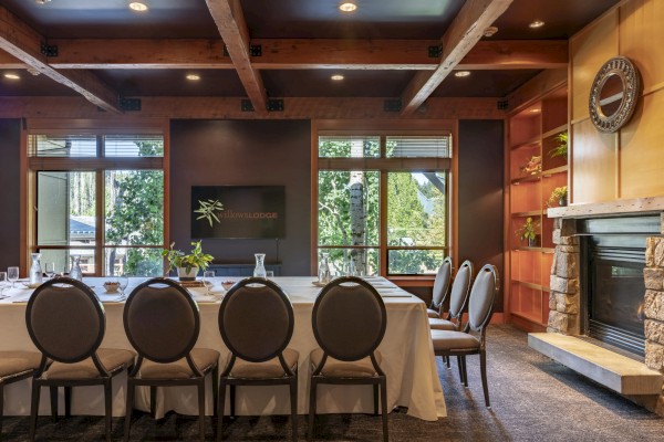 A cozy wooden meeting room with a long table set for a gathering, large windows, a fireplace, and a television displaying 
