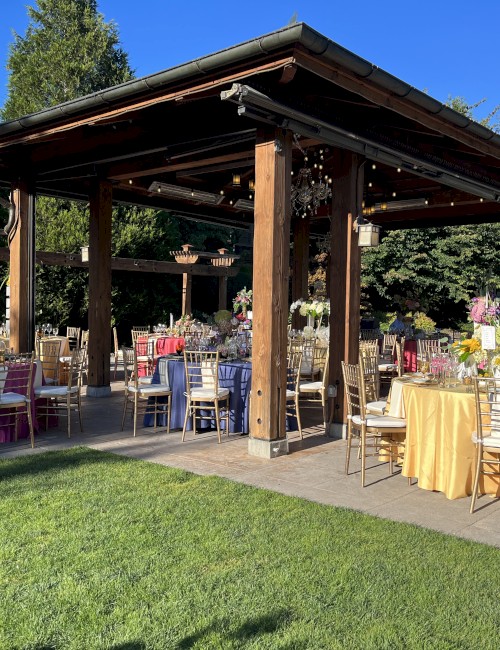 An outdoor event setup features a wooden pavilion, adorned with colorful table settings, chairs, and decor, surrounded by greenery under a clear blue sky.