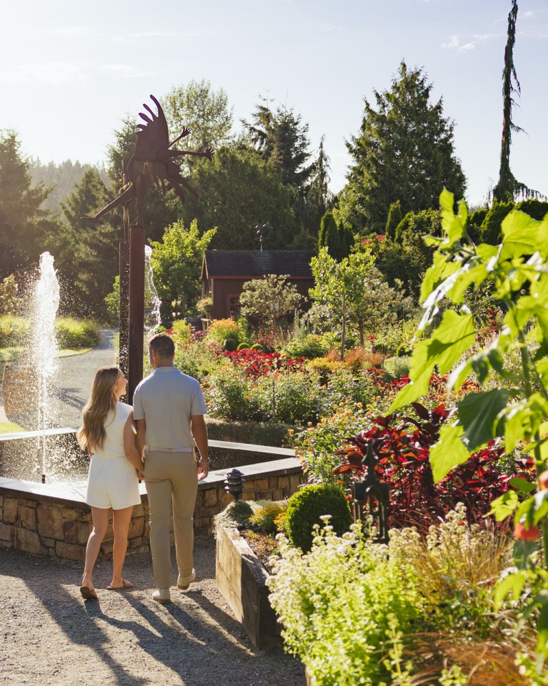 A couple walks hand-in-hand in a lush, colorful garden during sunny late afternoon or early evening.