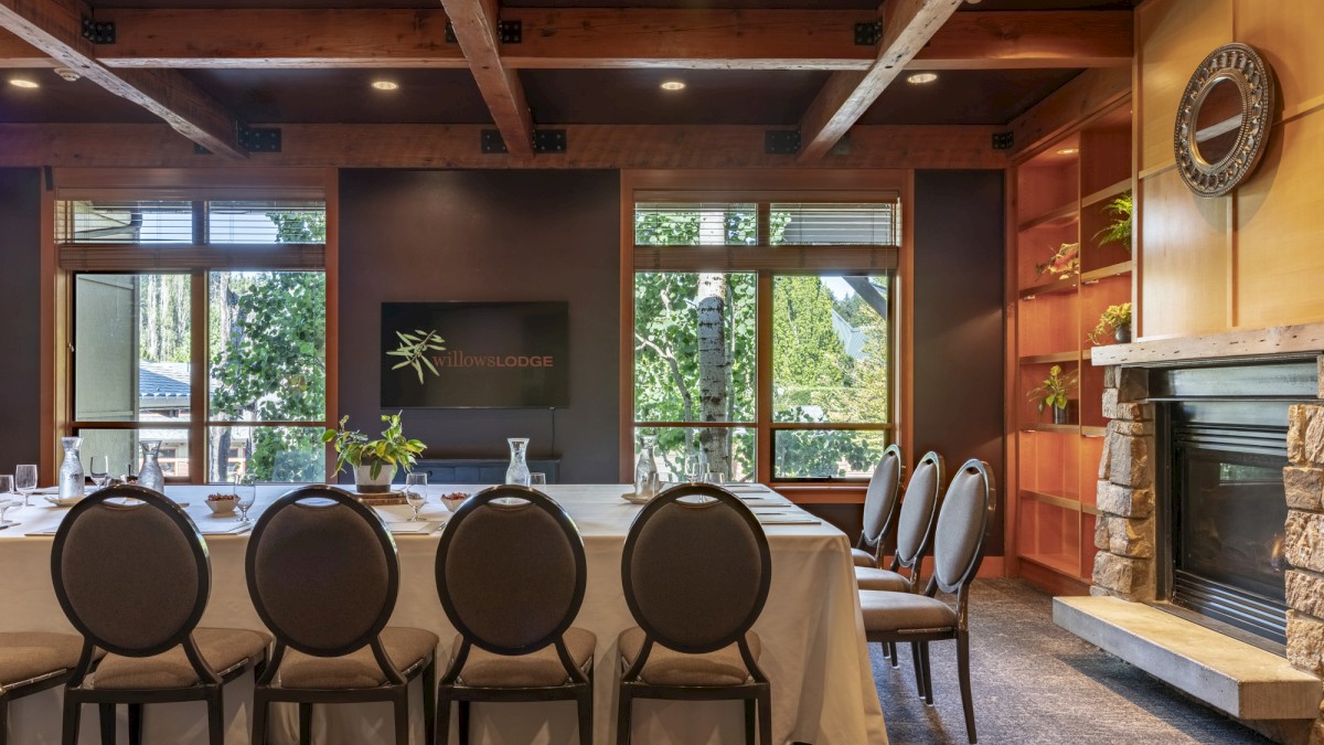 A cozy dining room with a long table set for a meeting or luncheon, surrounded by chairs, with a fireplace and large windows.