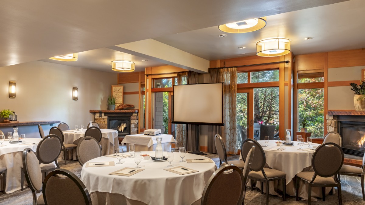 This image shows a conference room with round tables, place settings, and a projector screen, surrounded by windows with a garden view.