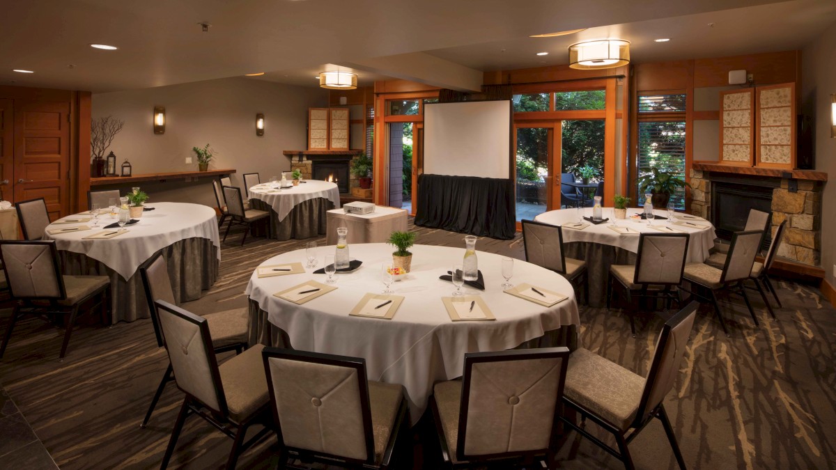A conference room setup with round tables, chairs, a projector screen, and notepads on tables, lit warmly with natural light from large windows.