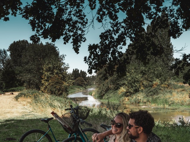 People enjoying a picnic outdoors under a large tree, near a river, with bikes, flowers, and drinks on a checkered tablecloth.