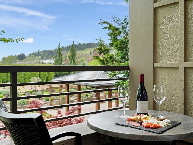 A balcony with a table set for wine and cheese, overlooking a lush garden and distant hills on a bright, clear day.