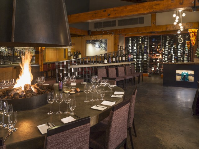 A cozy, upscale restaurant with a central fire pit, wine bottles on display, and an intimate ambiance with warm wooden decor.