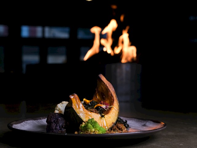 A gourmet dish with roasted pumpkin, colorful vegetables, and a fiery stove in the background.
