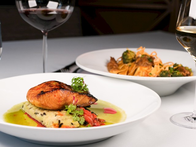 A gourmet meal with a grilled fish fillet served on a bed of vegetables, with side pasta, and glasses of white and red wine on a white table.