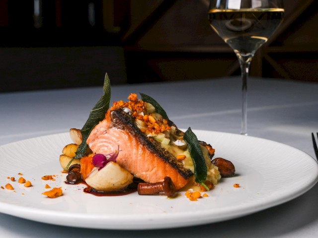 A beautifully plated piece of salmon with vegetables, garnished with herbs and served with a glass of white wine.