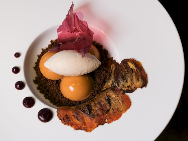 A plated dessert with chocolate crumble, mango gel, cream, a poached pear, a caramelized banana, and edible flower garnish.