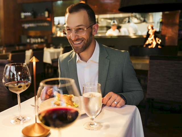 A man in glasses and a suit is enjoying a meal at a restaurant with glasses of wine and a slice of cake on the table.