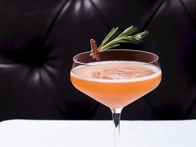 A pink cocktail garnished with a rosemary sprig on a white table against a black cushioned background.