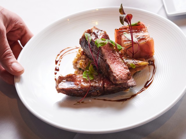 A hand holds a white plate with cooked meat, garnished with greens and drizzled with sauce, on a white table.