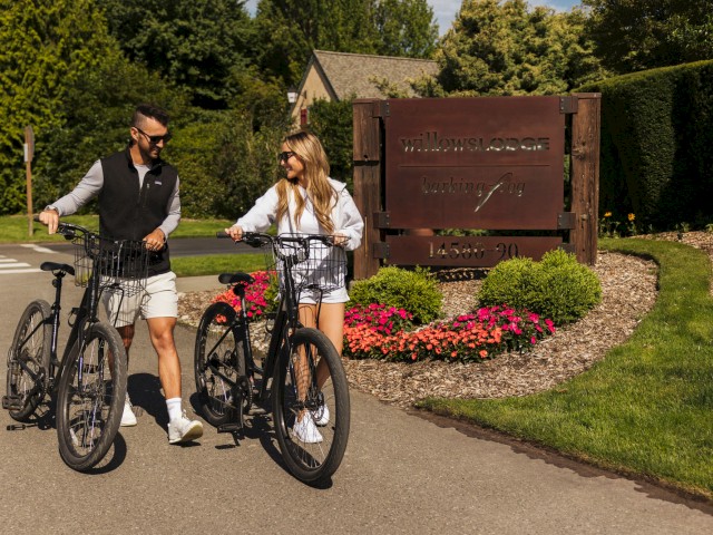 Two people with bikes are smiling and chatting near the entrance sign of Willows Lodge, a resort in a lush, green setting.