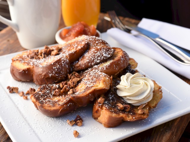 A plate of French toast topped with powdered sugar, crushed nuts, whipped cream, and served with syrup on the side.