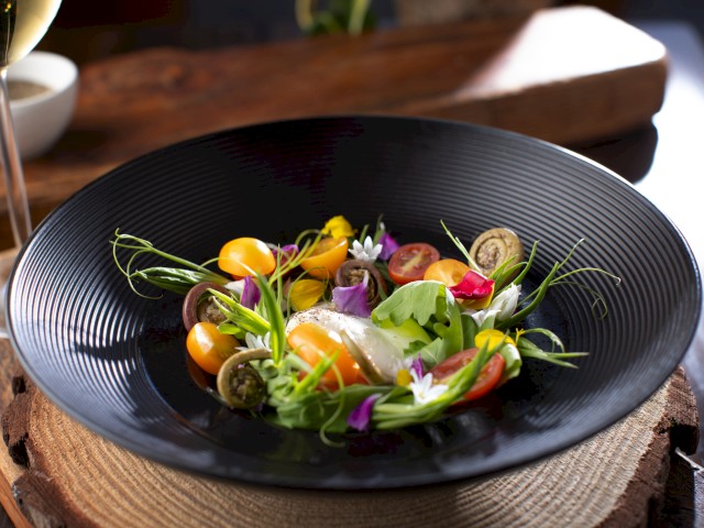 A colorful, elegantly plated salad with tomatoes, edible flowers, greens, and a poached egg on a black textured plate.