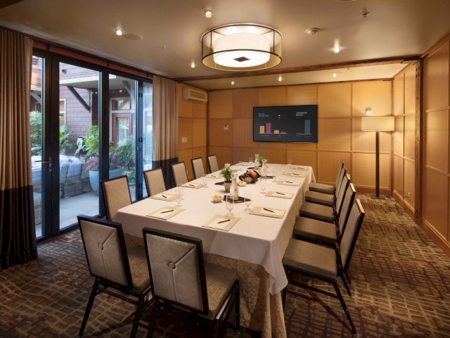 A meeting room set up for a formal event with a long table, chairs, a wall-mounted TV, and a standing lamp.