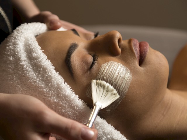 A woman receives a facial treatment where a skincare product is being gently applied with a brush, while she rests with closed eyes.