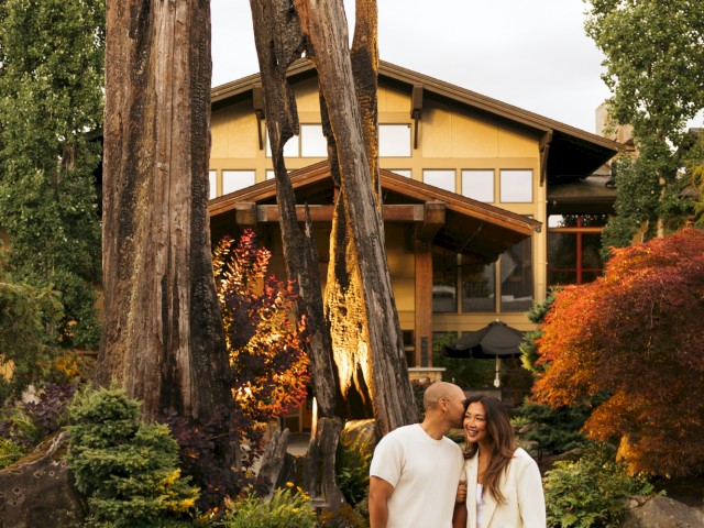A couple walks happily in front of a house with tall, charred wood sculptures and lush greenery in the background.